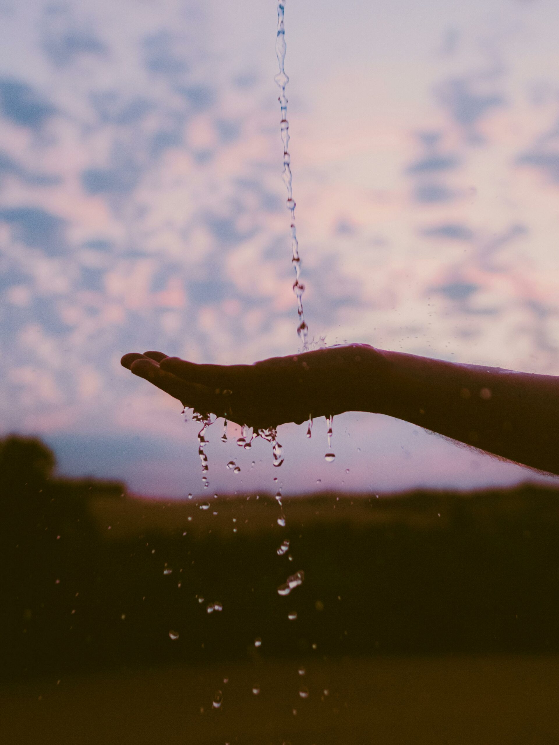 Une main tendue récupérant des goutes d'eau, devant un ciel mauve, en début de soirée